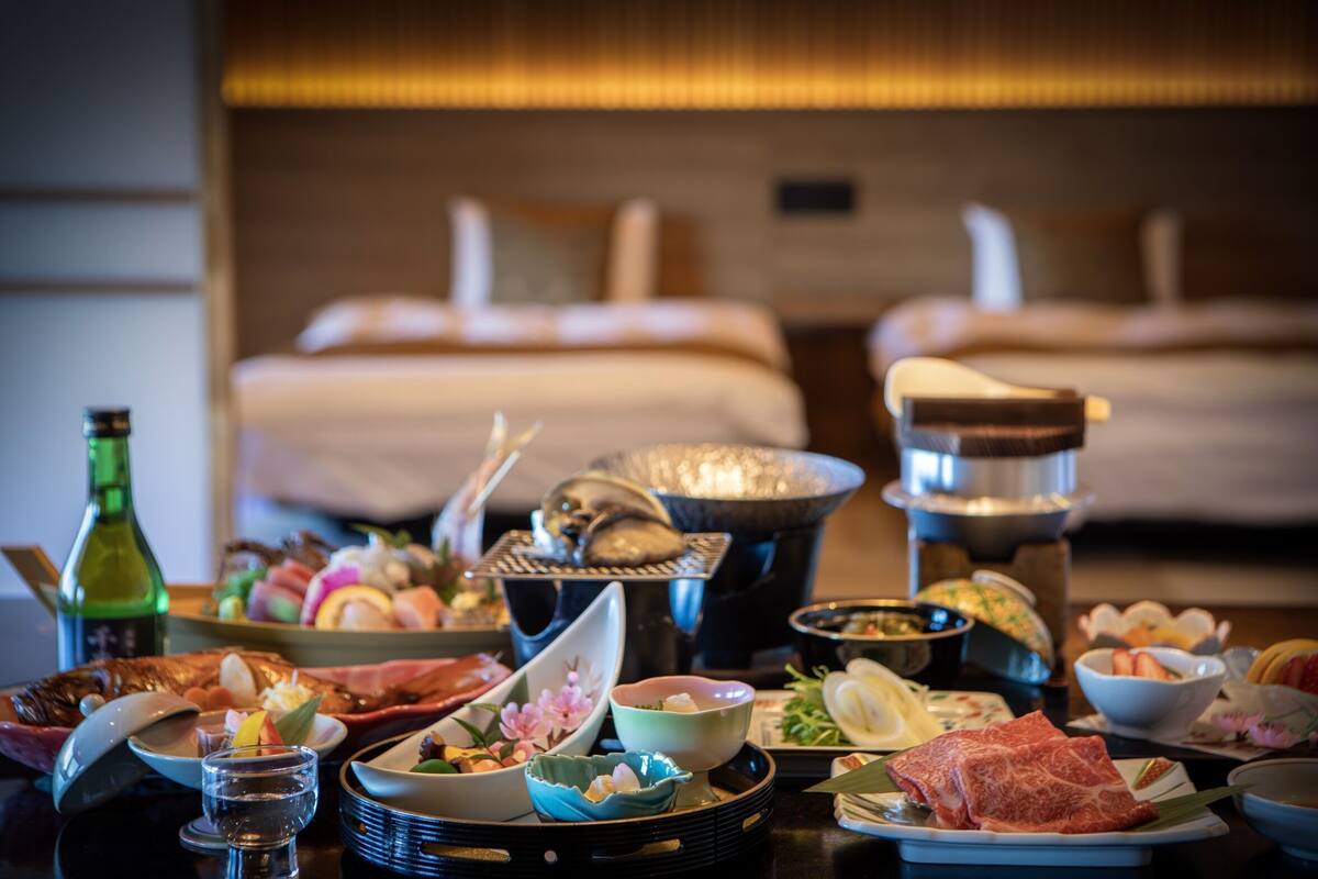 A dining table with various dishes and a hot pot, a bottle, and a bed in the background.