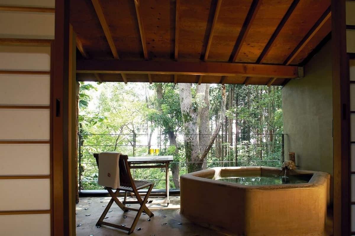 Ein traditioneller japanischer Raum mit Tatami-Böden, einer Holzdecke, einer Onsen-Badewanne und Blick auf einen Wald.