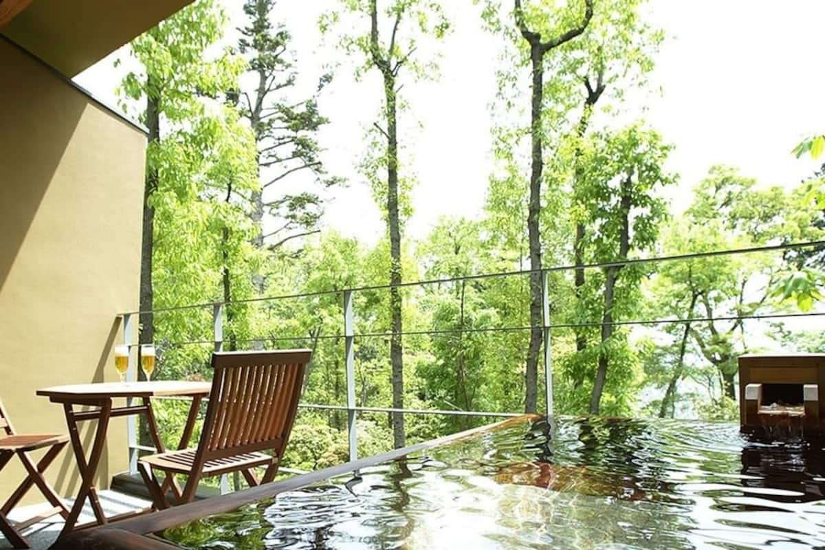 Ein Innenpool mit Infinity-Kante, umgeben von Holenstühlen und einem Tisch, mit Glaswänden und Blick auf Bäume.