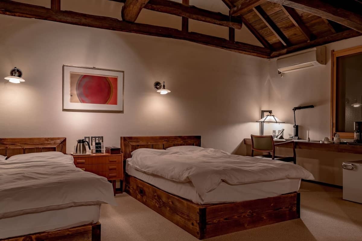 Ein Hotelzimmer mit zwei hölzernen Betten, einem Schreibtisch mit Stuhl, einer Schreibtischlampe, einer Kaffeemaschine und einem großen Spiegel.