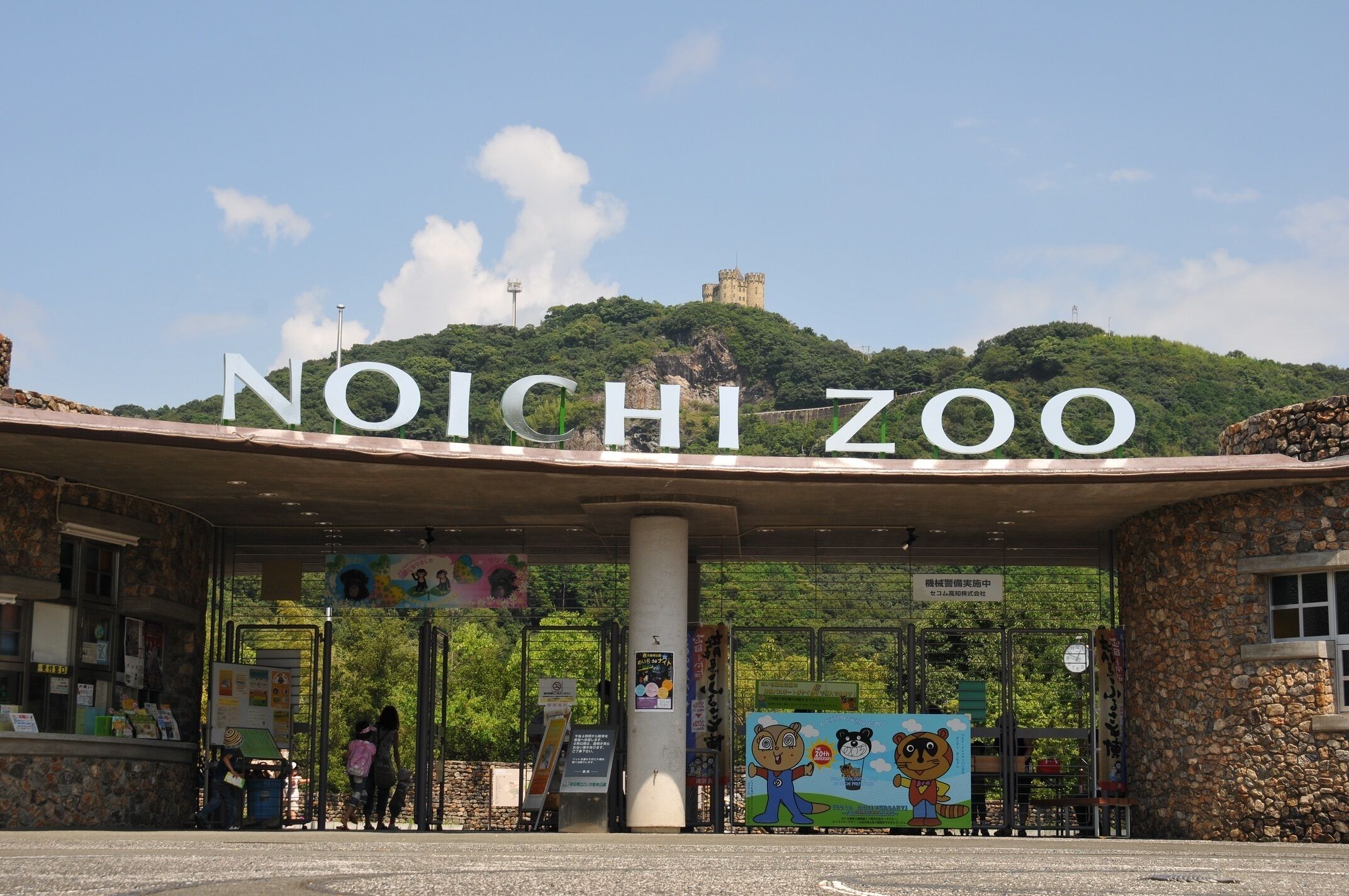 De ingang van Noichizoo met een groot bord, een stenen gebouw en een uitzicht op een kasteel op een heuvel.