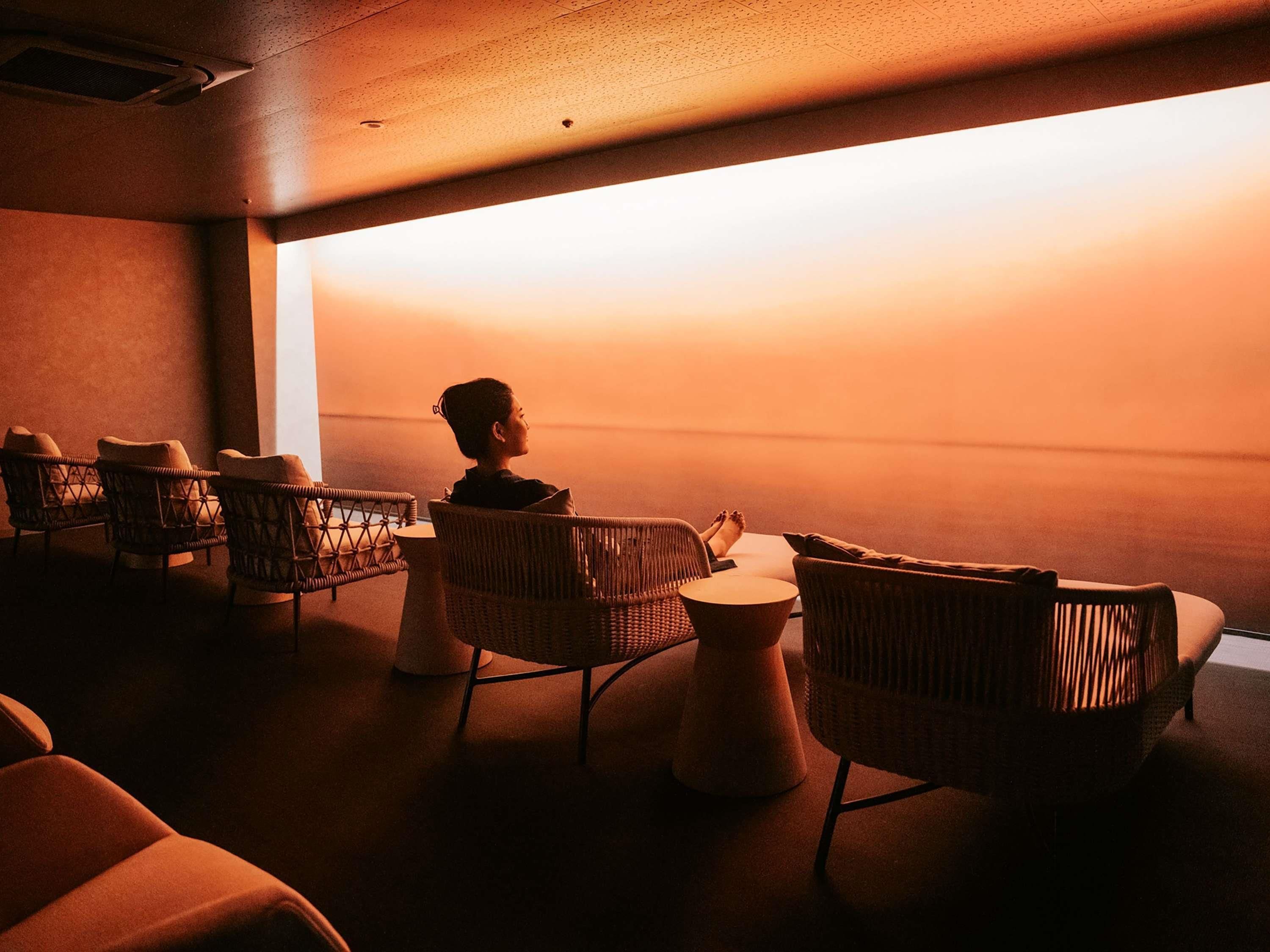 A room with wicker chairs and a small round table, a person sitting by a large window, and a ceiling vent.
