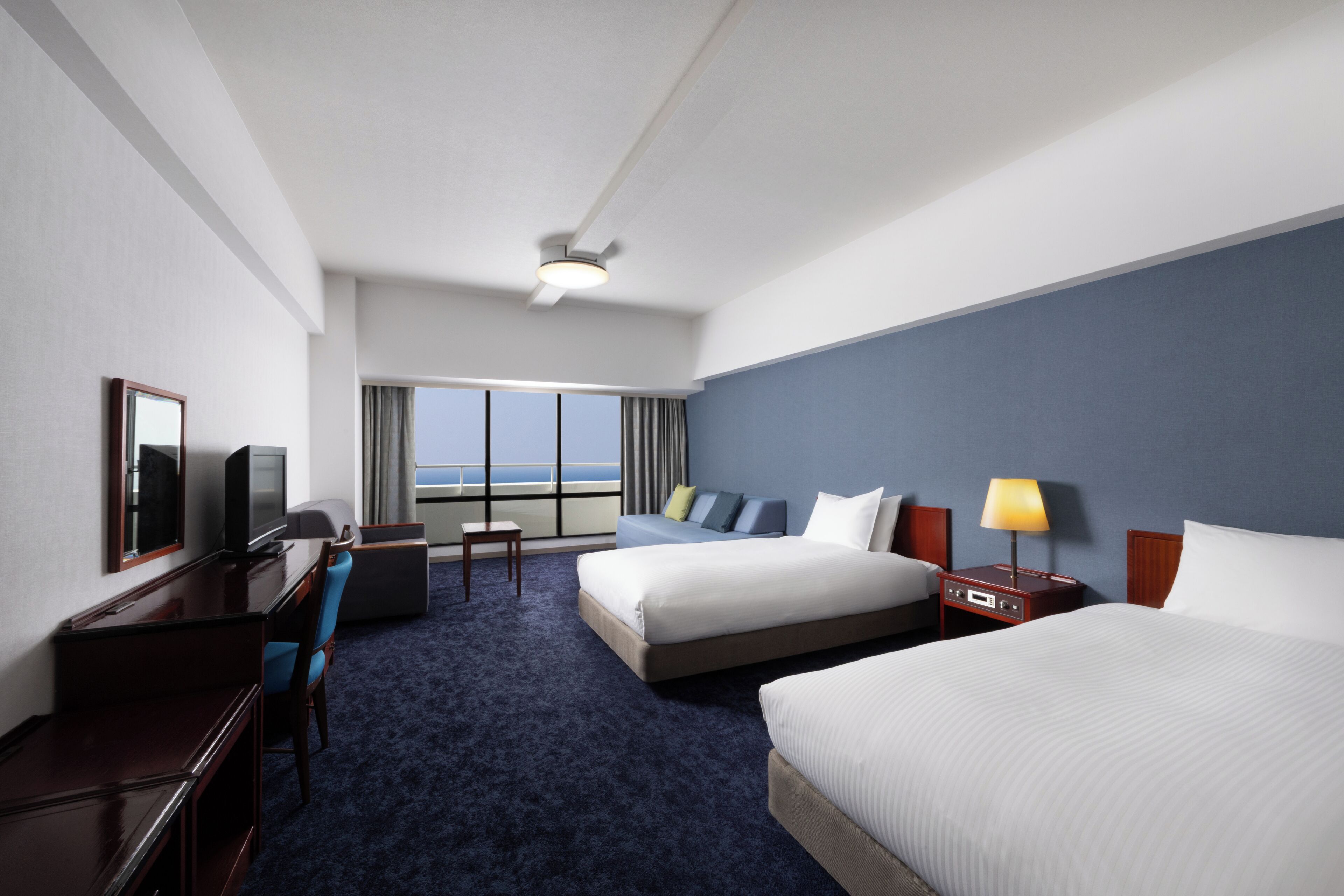 Una habitación de hotel con una cama grande, una cama más pequeña, un escritorio con una silla, un televisor y una ventana con vista al océano.