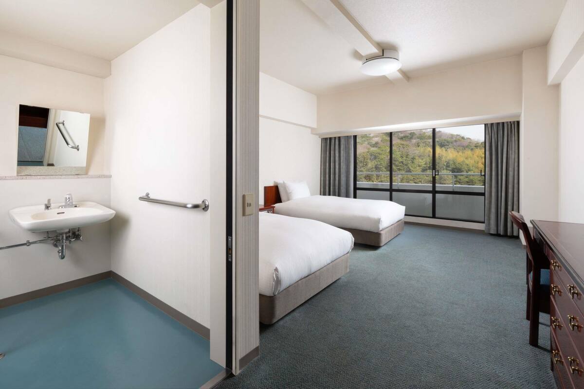 A hotel room with a bathroom, two beds, a desk, and a large window with a view.