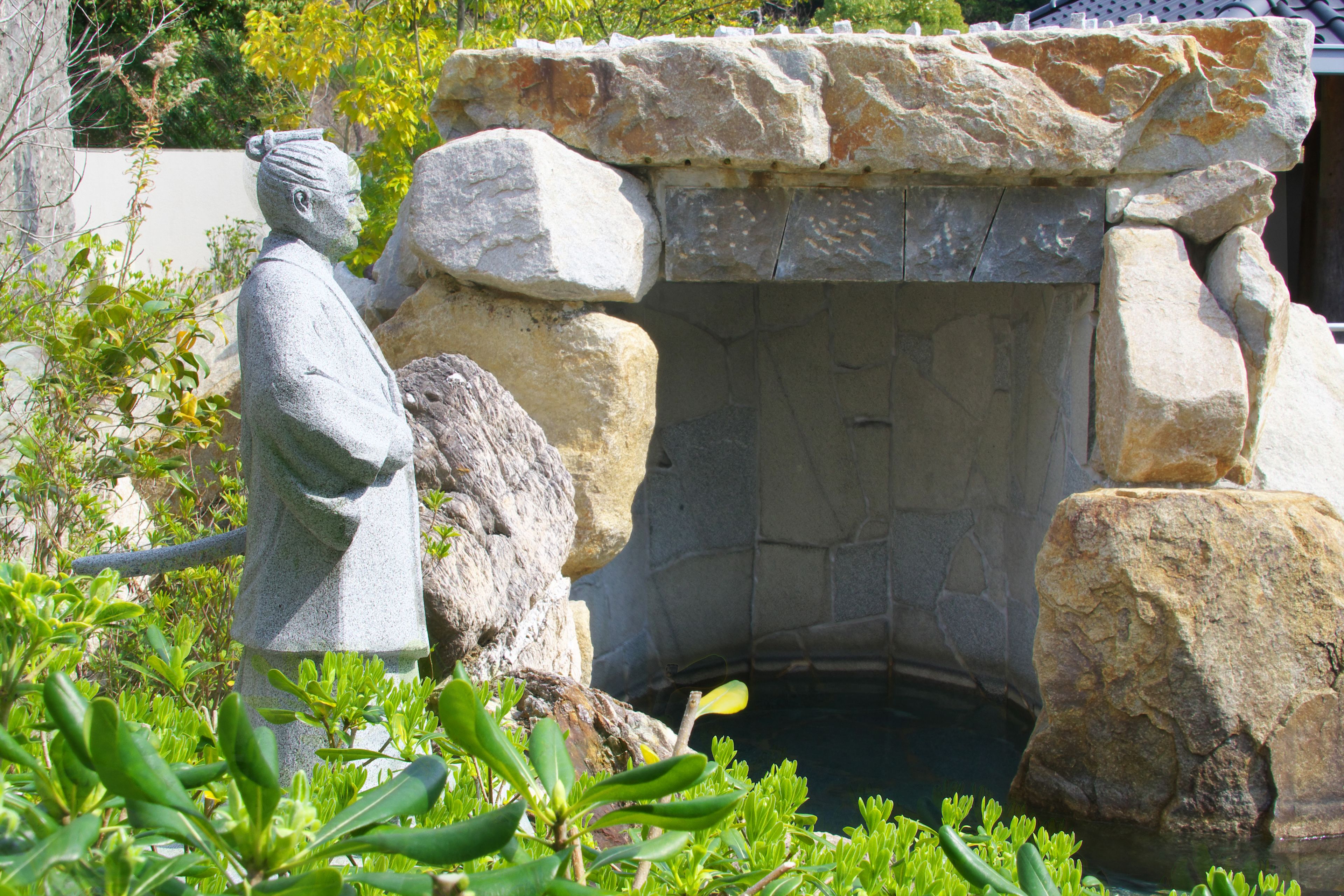 A stone statue of a figure in a garden setting with a rock structure and greenery.