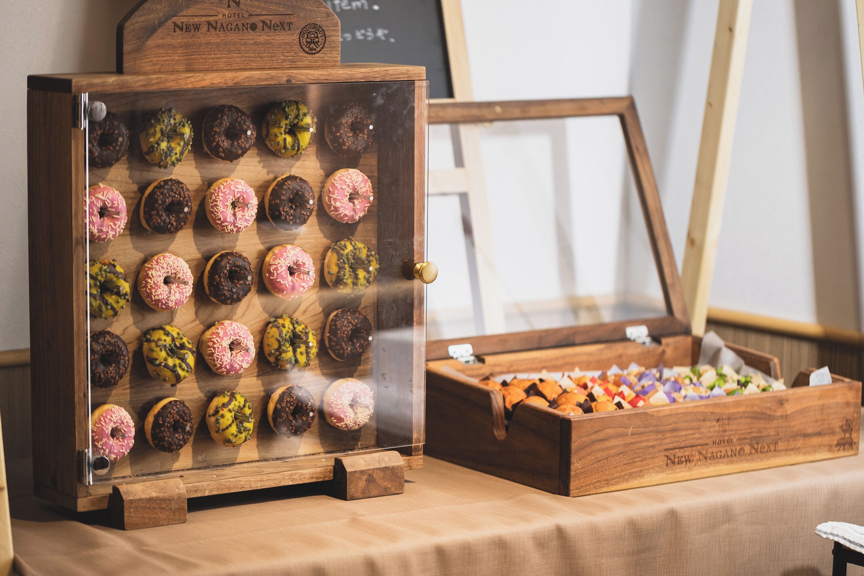 Uma vitrine de madeira com donuts variados e uma caixa de madeira com uma placa que diz 'New Nagano Next Hotel'.