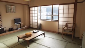 Una habitación japonesa tradicional con suelo de tatami, una mesa de madera, un televisor sobre un soporte y vistas a una masa de agua a través de paneles shoji.