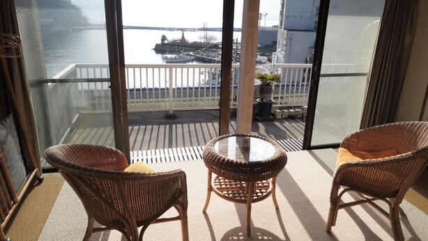 Un balcón con sillas de mimbre y una pequeña mesa, con vistas a un puerto deportivo con barcos y un puente.