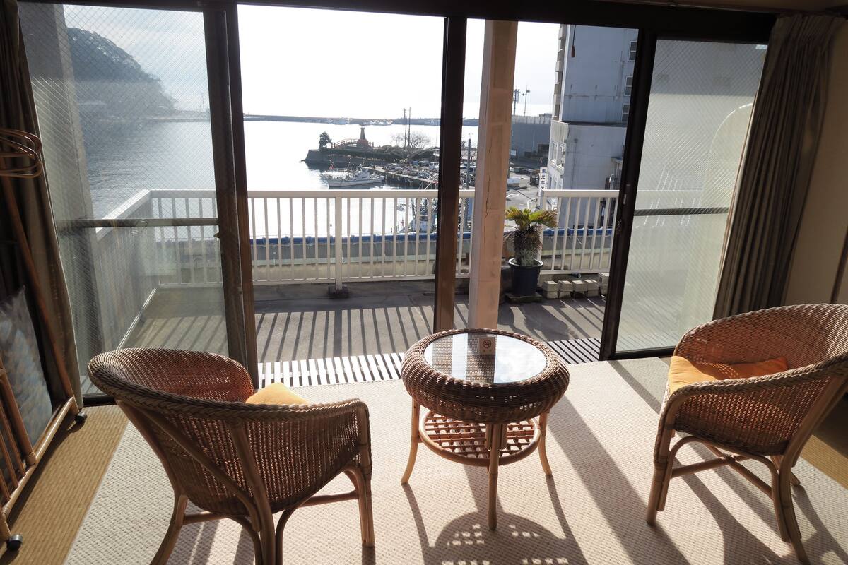 A balcony with wicker chairs and a small table, overlooking a marina with boats and a bridge.