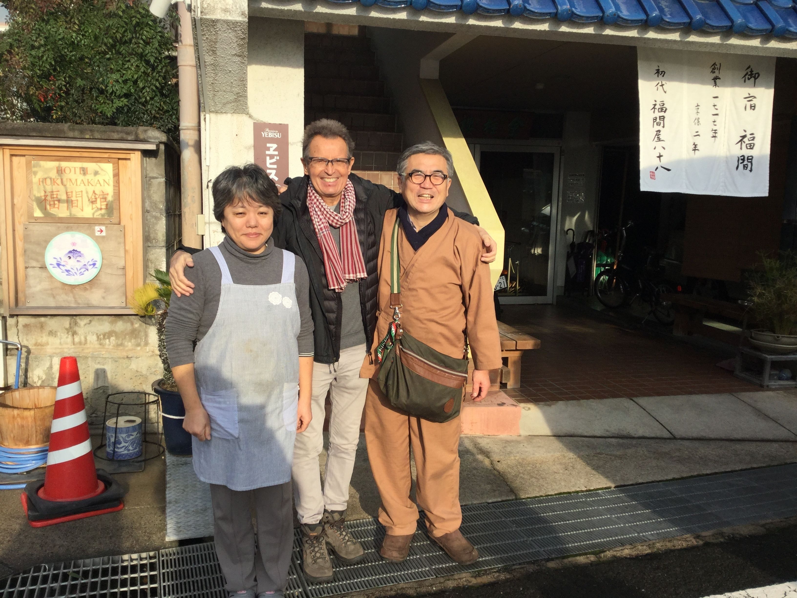 Three people standing in front of a building with a signboard and a wooden plaque.