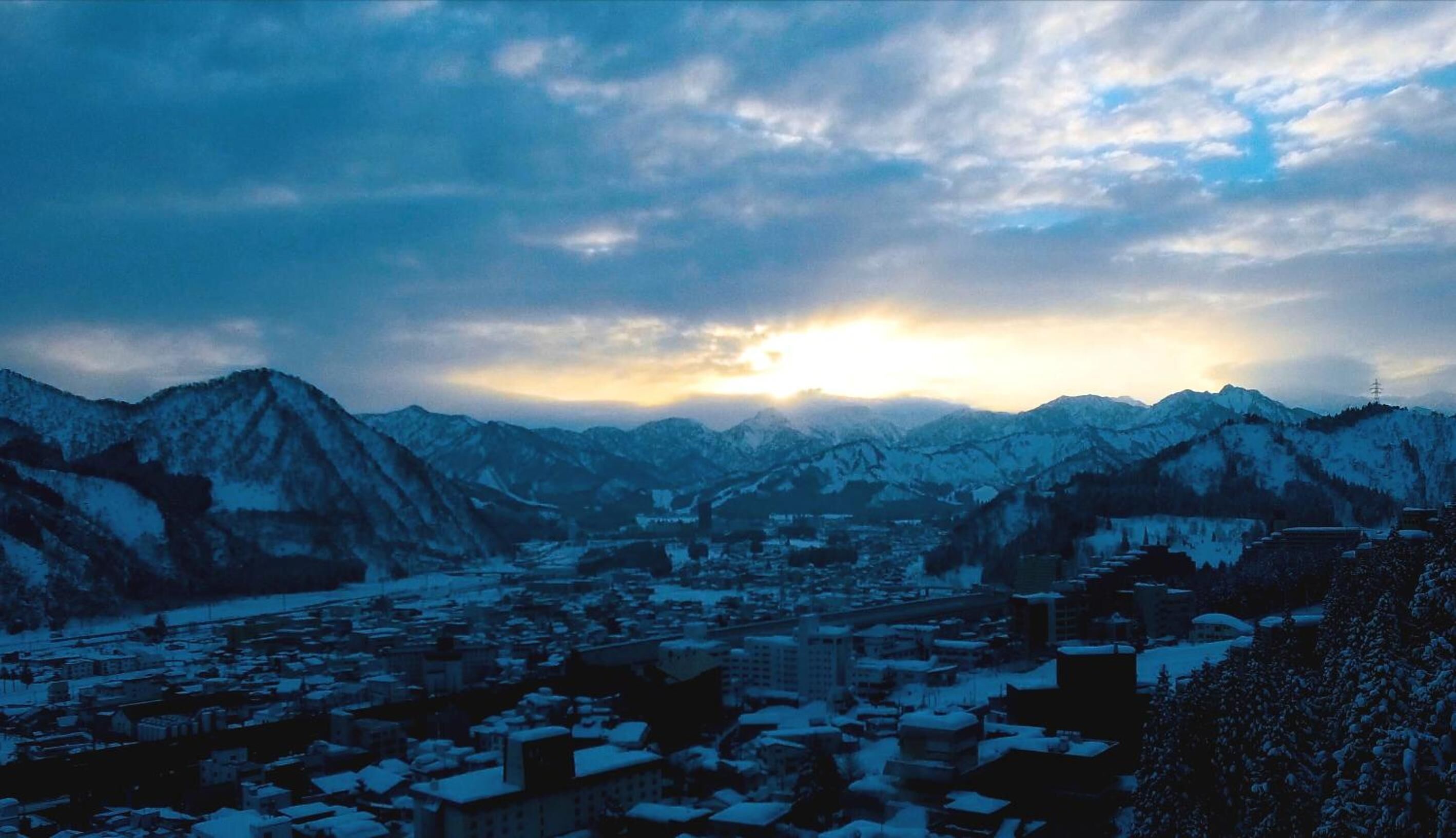 A town nestled between snow-covered mountains with a clear sky and the sun setting in the background.