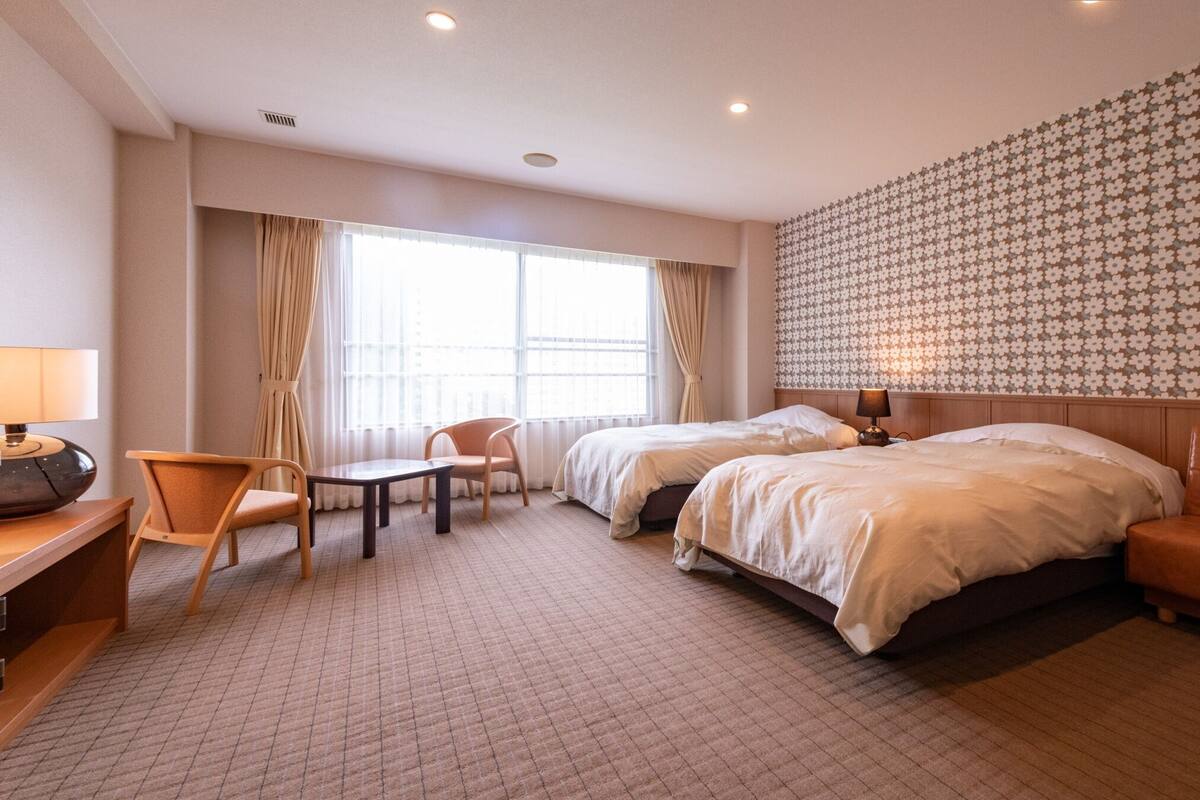A hotel room with two beds, a desk, a chair, a lamp, and a patterned wall.