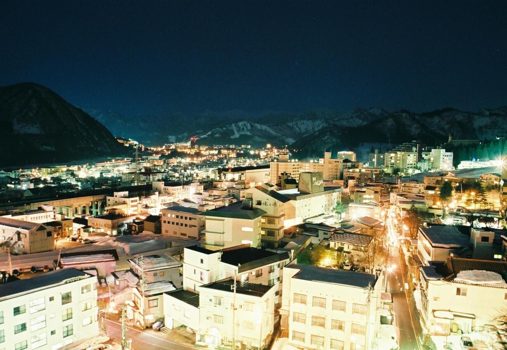 A nighttime cityscape with buildings, roads, and mountains in the background.