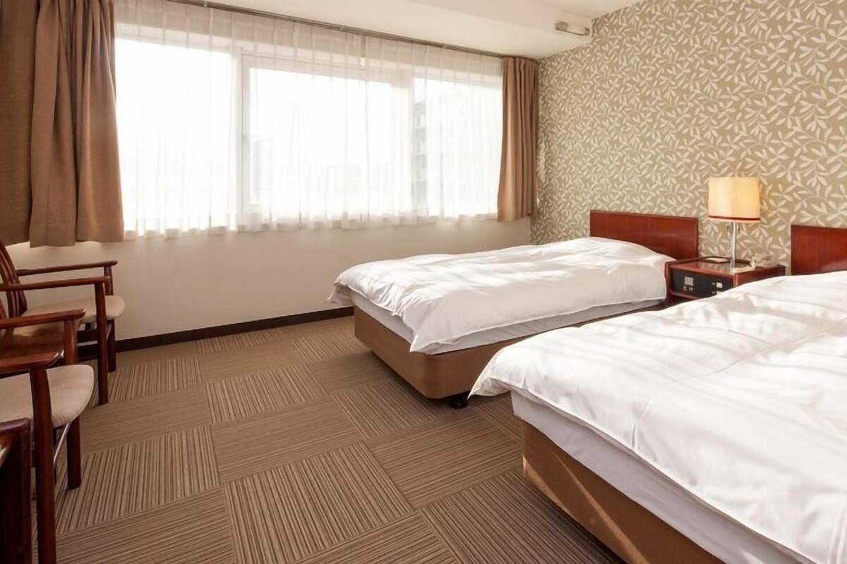 A hotel room with two beds, a desk, a chair, a lamp, and a patterned wall.