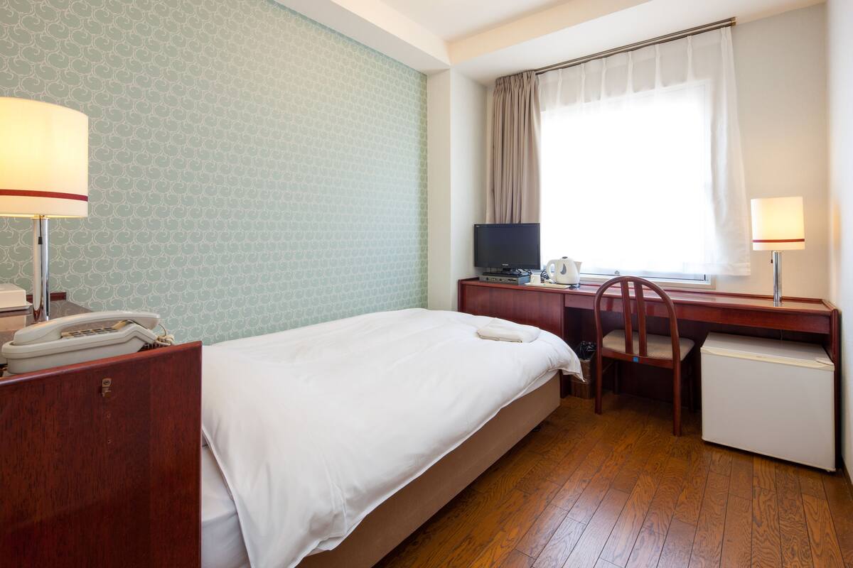 A hotel room with a bed, desk, chair, and a small refrigerator.