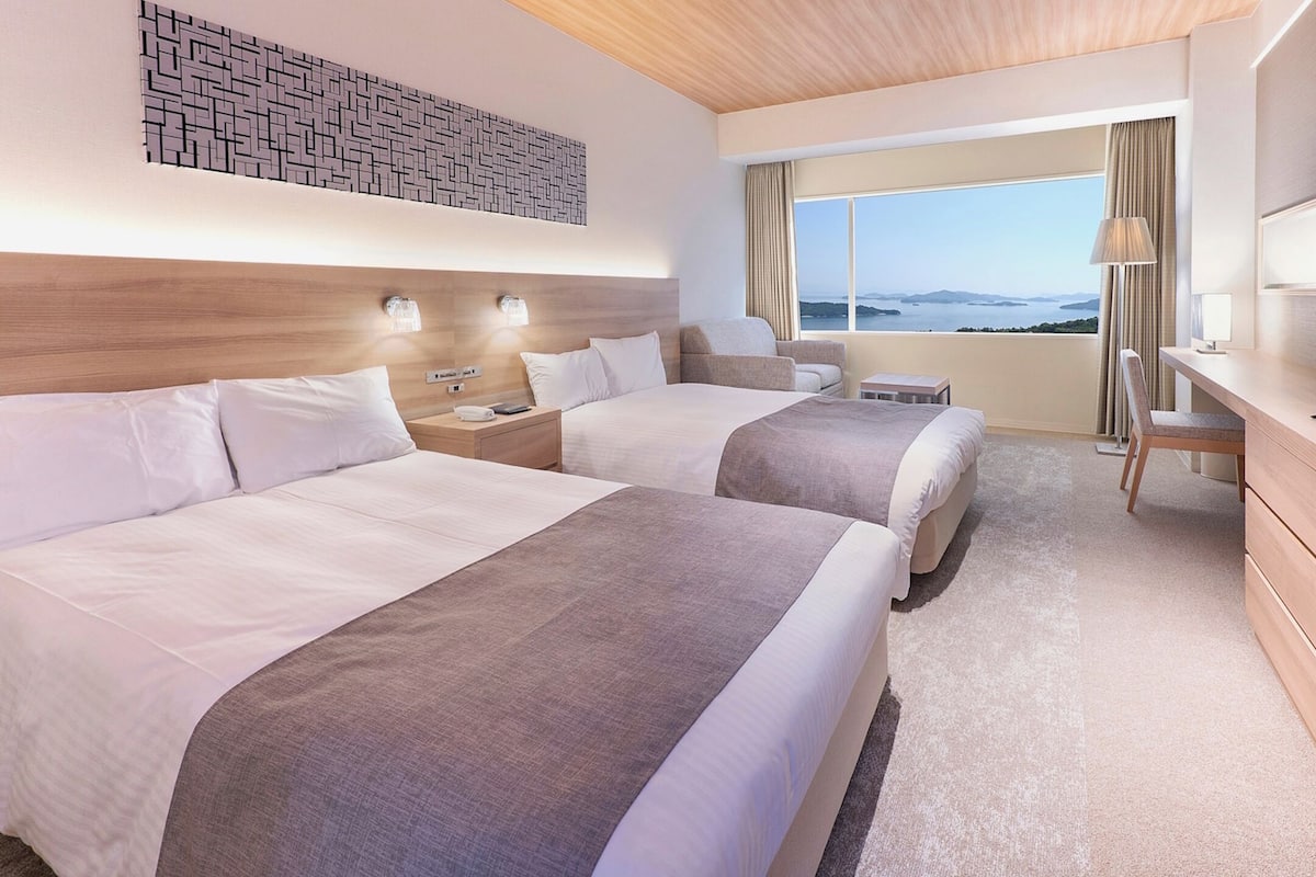 A hotel room with two beds, a TV, a desk, and a view of the sea.