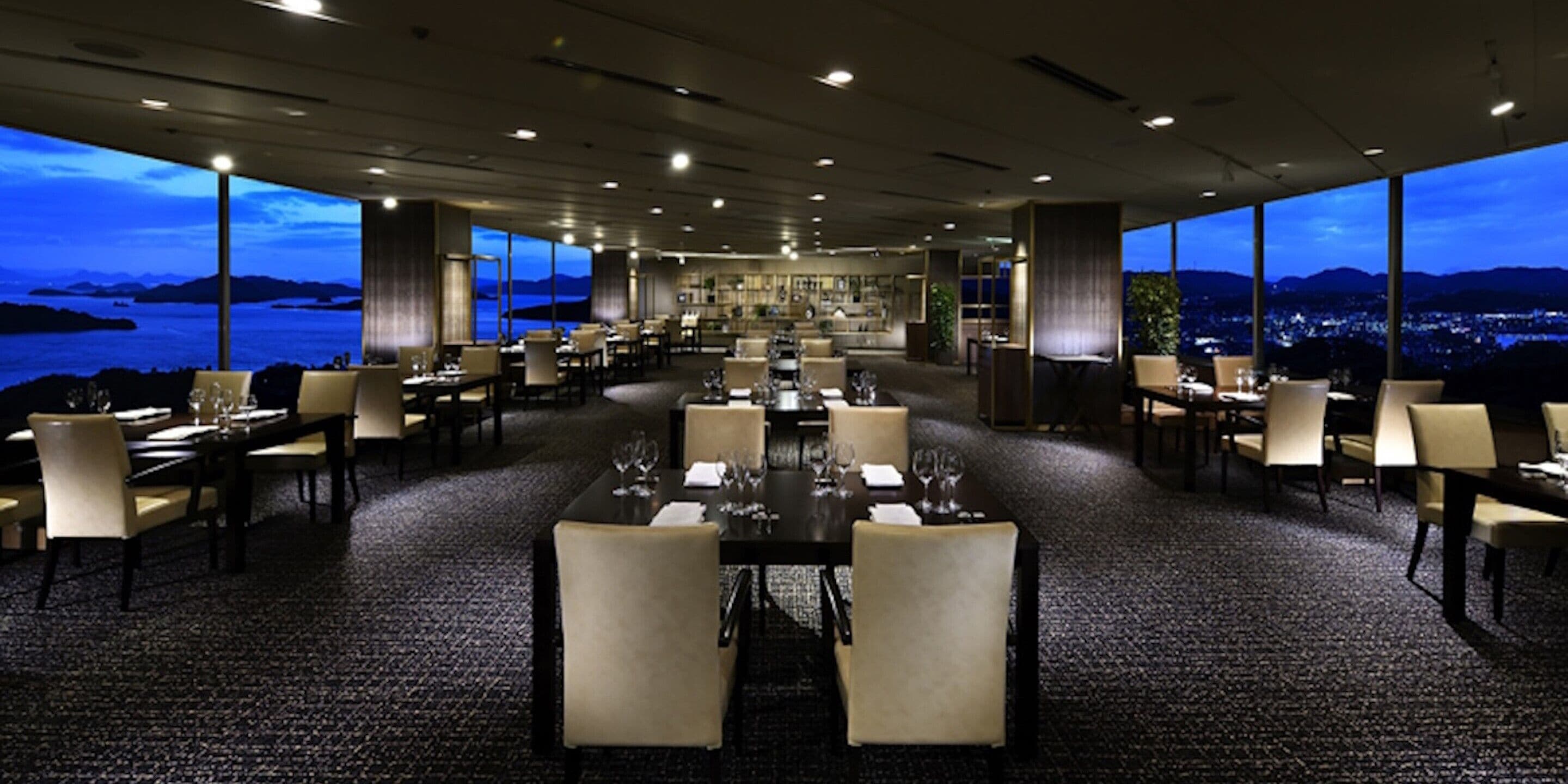 Un comedor espacioso con múltiples mesas y sillas, amplias ventanas que ofrecen vistas al mar y al paisaje urbano, y un diseño interior moderno.