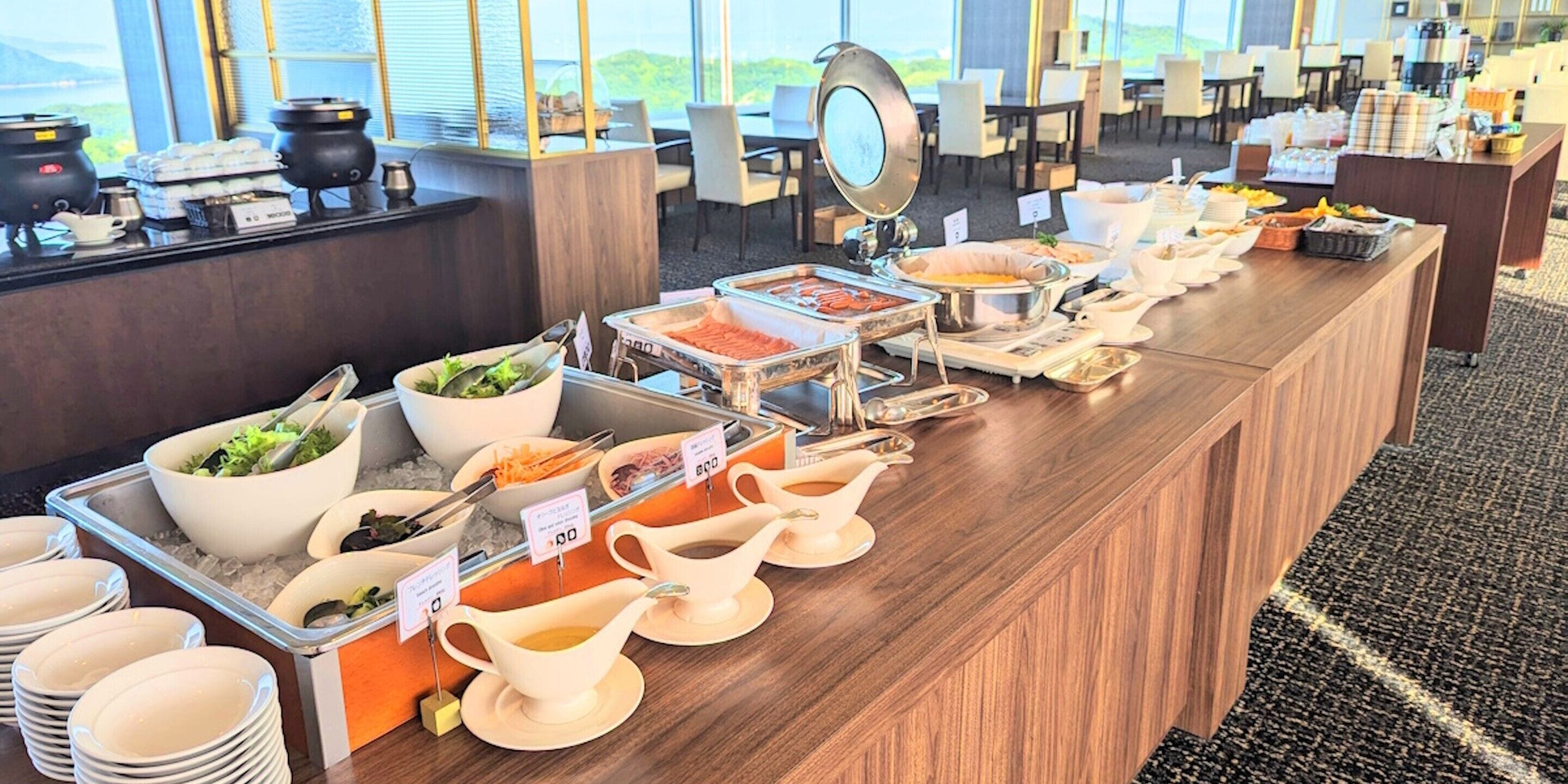 A buffet setup with various food stations, including salads, hot dishes, and beverages, in a well-lit room with a view of the outdoors.