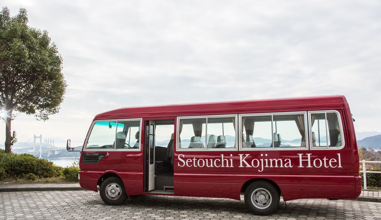 Ein roter Bus mit der Aufschrift "Setouchi Kojima Hotel" auf der Seite, geparkt auf einem Kopfsteinpflasterweg mit einer Brücke und Grünflächen im Hintergrund.