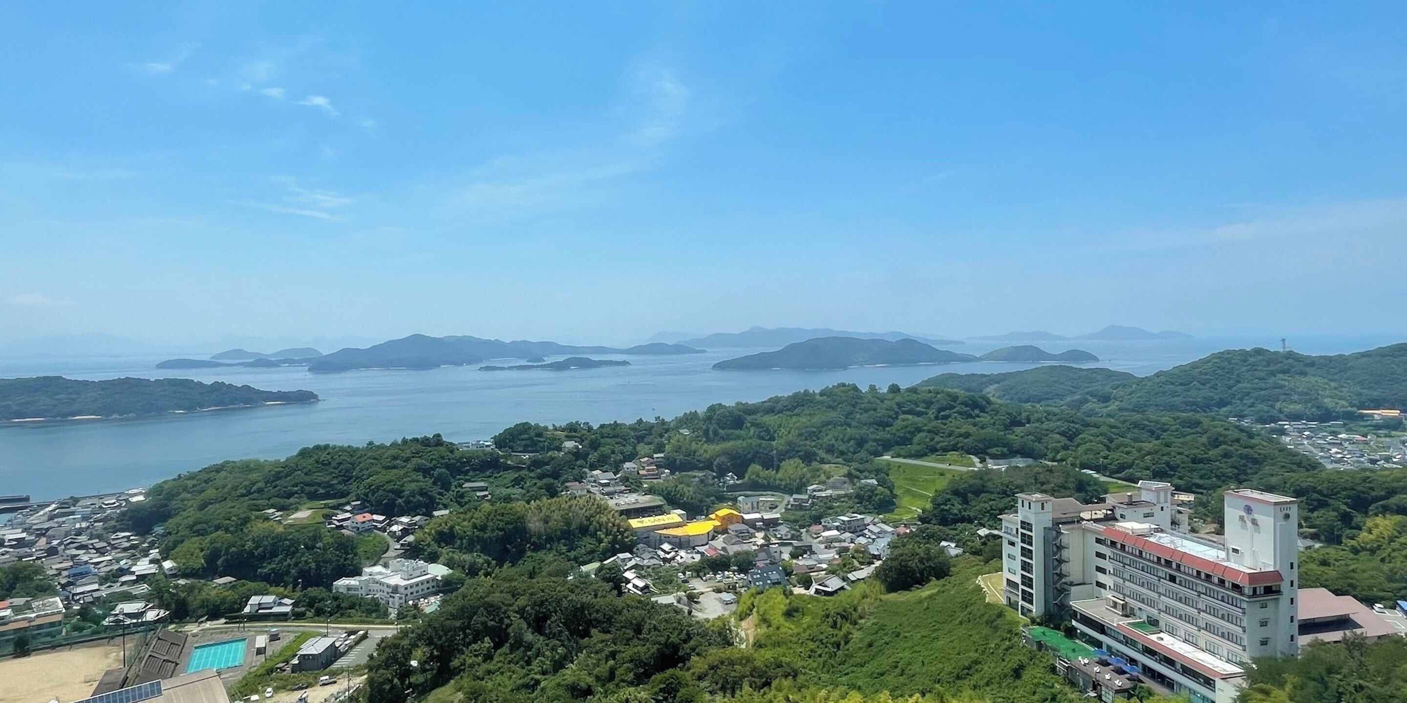 A coastal town with a prominent building featuring a red roof and a white tower, surrounded by lush greenery and a clear blue sky.