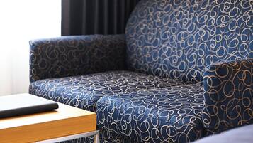 Un divano angolare con un motivo blu e bianco, un tavolino in legno con un oggetto nero sopra e una tenda scura sullo sfondo.