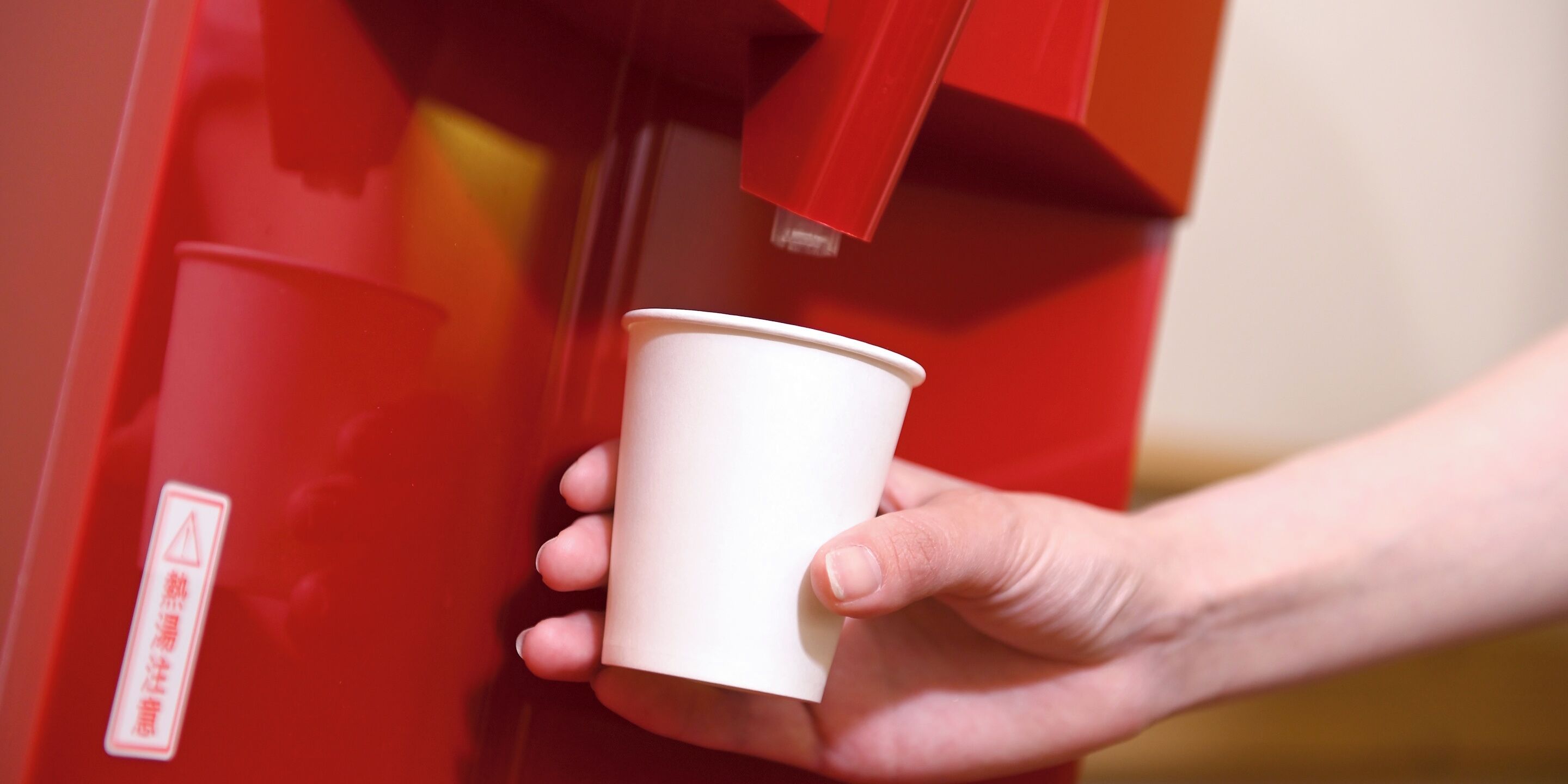 Una mano sostiene una taza blanca cerca de una máquina expendedora roja.