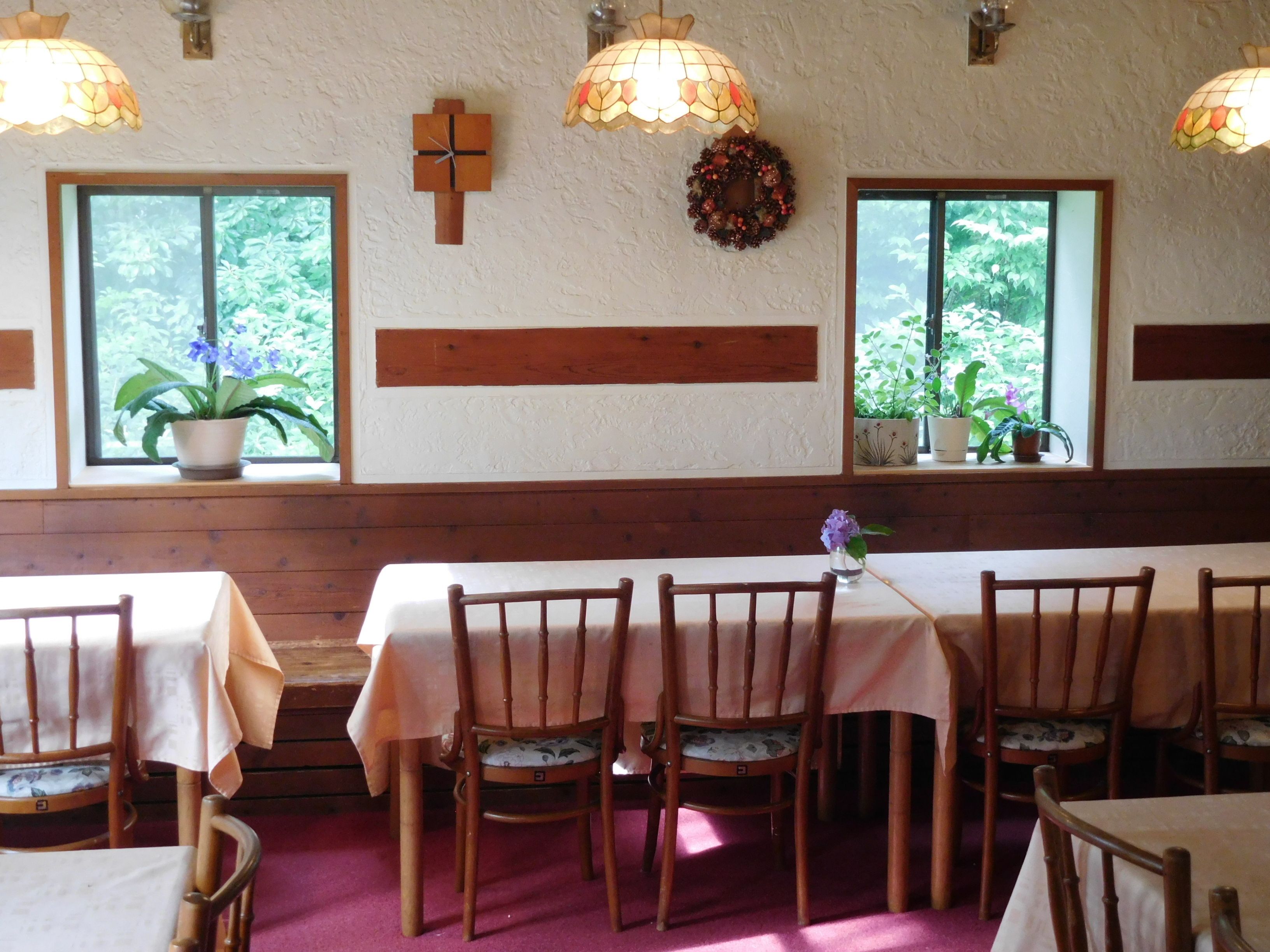 A dining area with wooden tables and chairs, a cross on the wall, and windows with plants.