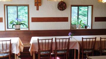 A dining area with wooden tables and chairs, a cross on the wall, and windows with plants.