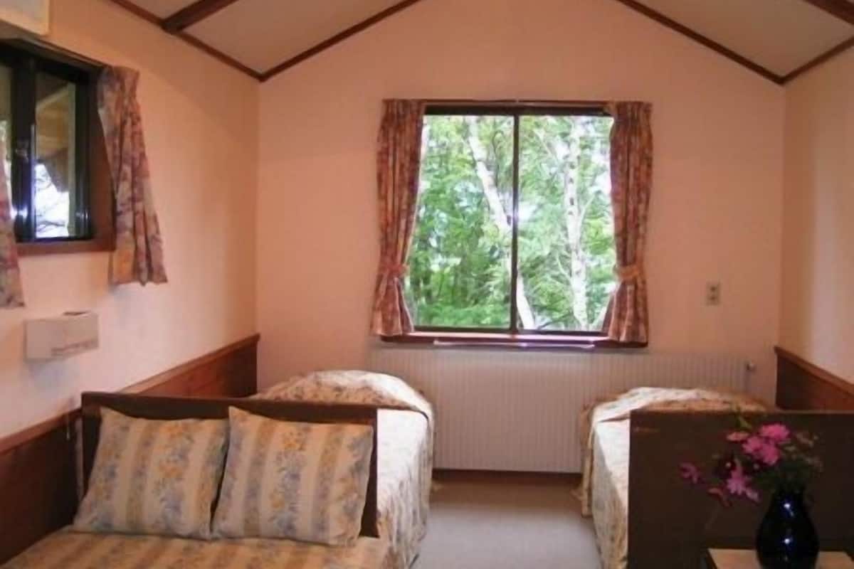 Quarto com duas camas, janela com cortinas, luminária no teto e um vaso com flores sobre uma mesa.