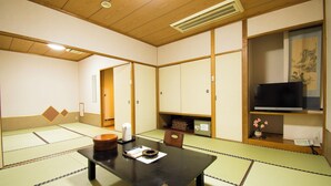 Um quarto com tatames, uma mesa, cadeiras, aquecedor, televisão e arte japonesa tradicional.