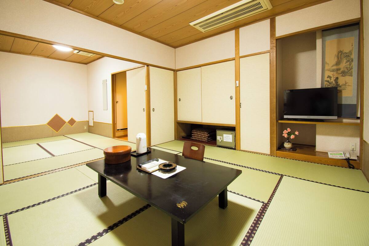 A tatami mat room with a table, chairs, a heater, a TV, and traditional Japanese artwork.