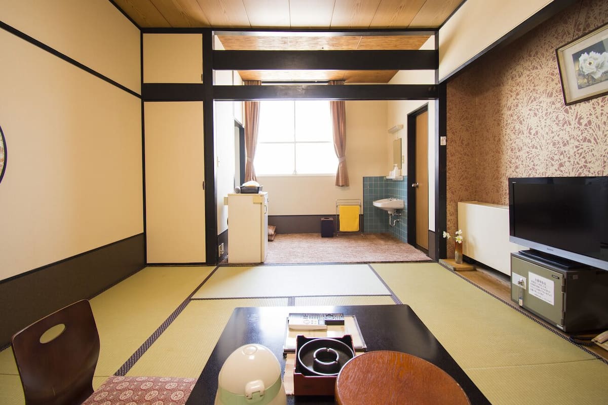 A traditional Japanese-style room with tatami flooring, a television, a small refrigerator, and a sliding door leading to a bathroom.