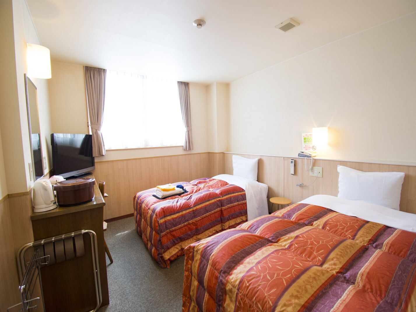Ein Hotelzimmer mit zwei Betten, einem Fernseher, einem Schreibtisch mit Lampe und einem Fenster mit Vorhängen.