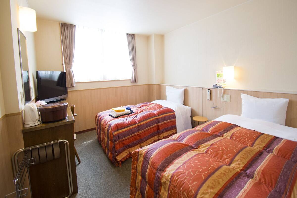 A hotel room with two beds, a TV, a desk with a lamp, and a window with curtains.