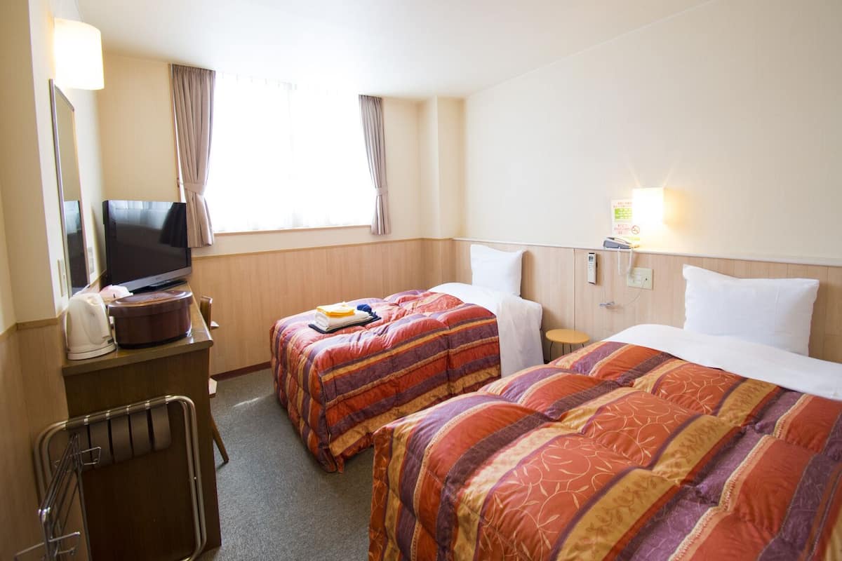 Ein Hotelzimmer mit zwei Betten, einem Fernseher, einem Schreibtisch mit Lampe und einem Fenster mit Vorhängen.