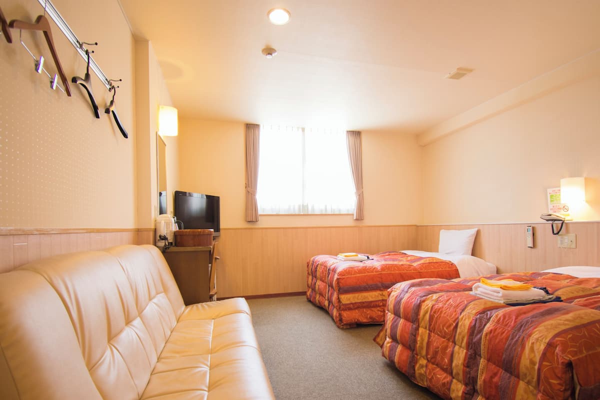 A hotel room with a sofa, two beds with colorful bedding, a television, and a window with curtains.