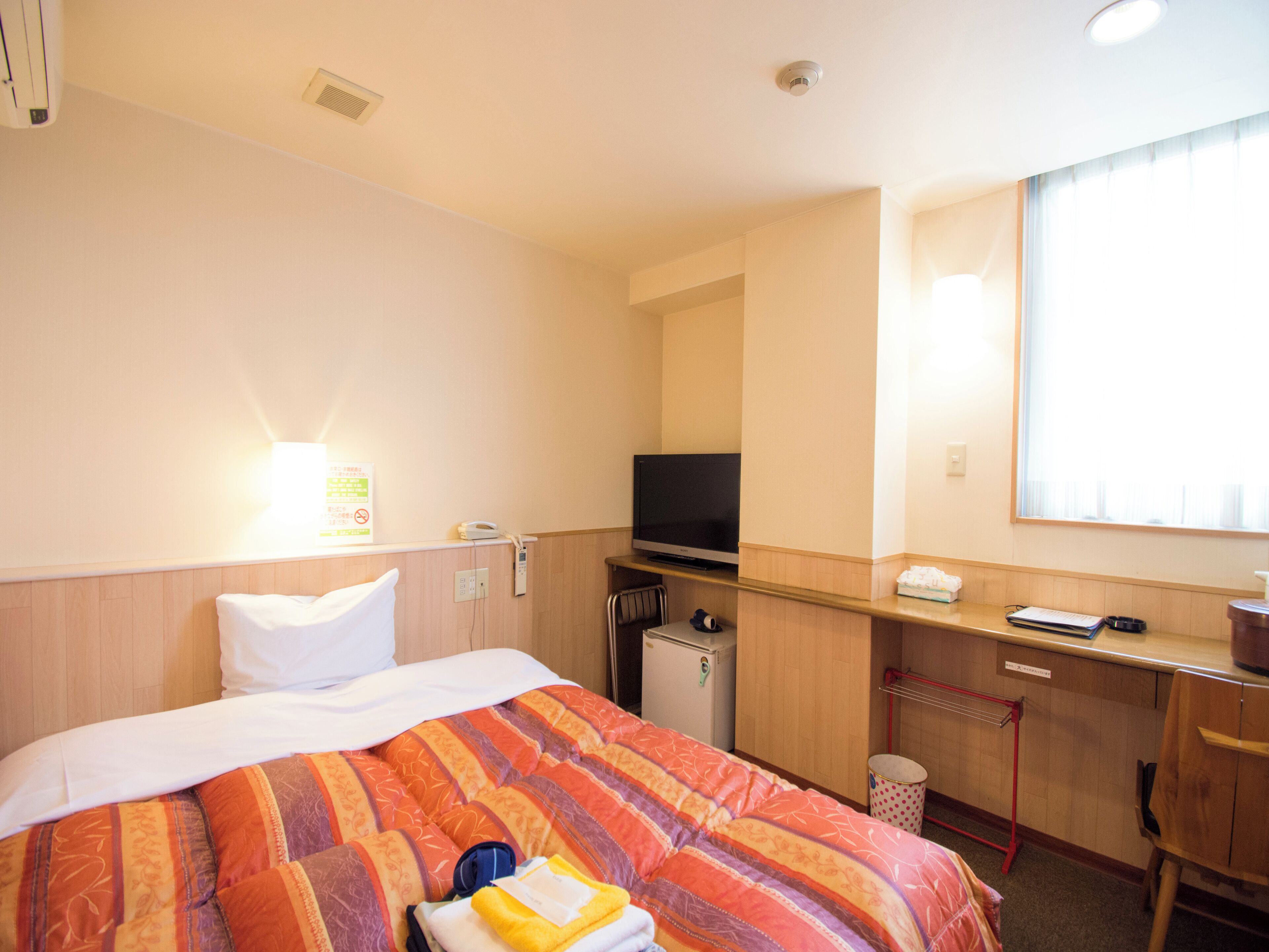 A hotel room with a bed, a desk with a television, a small refrigerator, and a window with blinds.