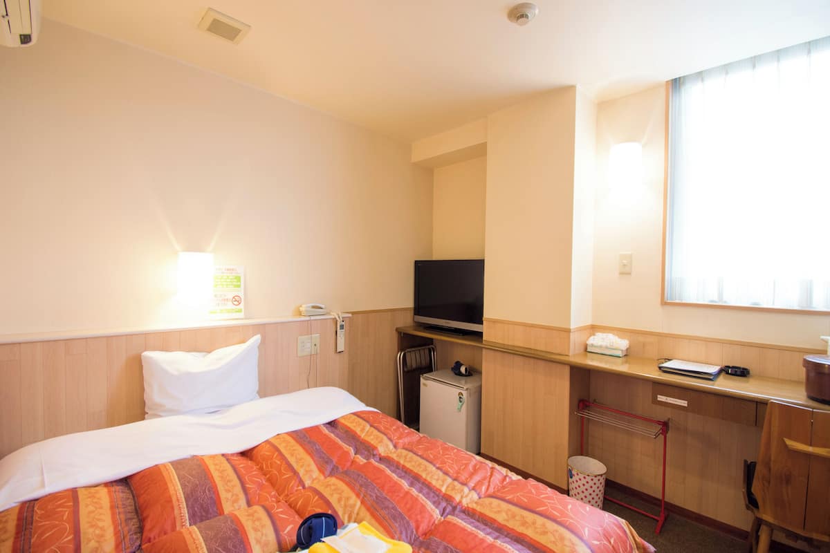 A hotel room with a bed, a desk with a television, a small refrigerator, and a window with blinds.