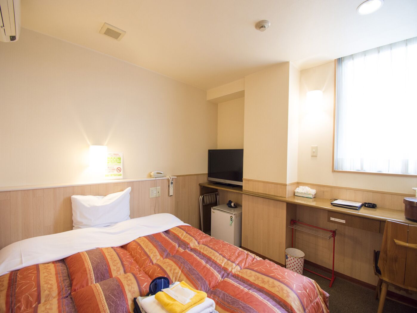 Ein Hotelzimmer mit einem Bett, einem Schreibtisch mit Fernseher, einem kleinen Kühlschrank und einem Fenster mit Jalousien.