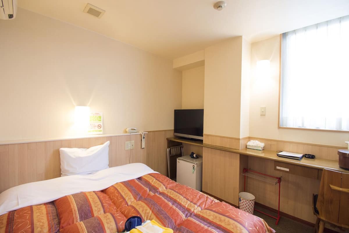 A hotel room with a bed, a desk with a television, a small refrigerator, and a window with blinds.