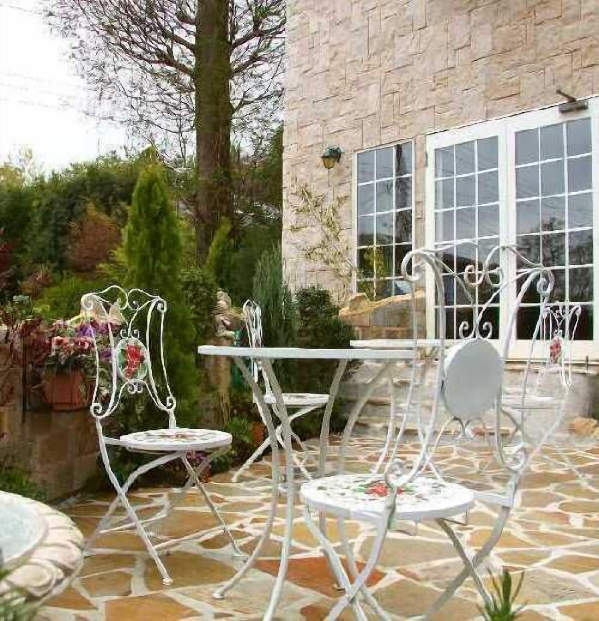 Un patio con sillas y mesa de metal blanco, rodeado de piso de piedra y follaje.