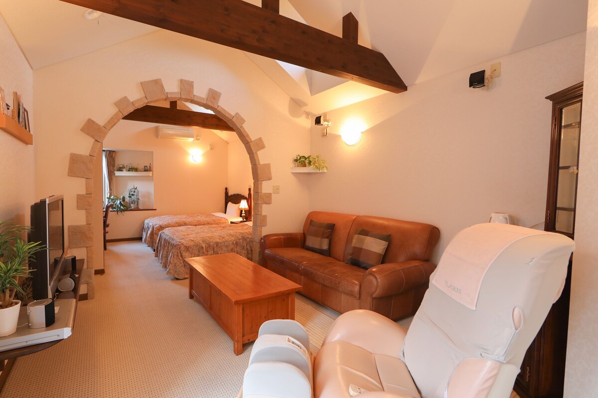 A cozy room with a wooden bench, a leather sofa, and a white armchair. There is a bed with a wooden headboard and a small table with a lamp.