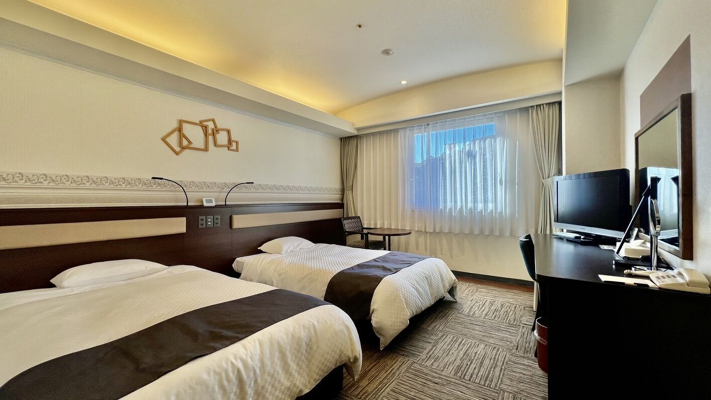 Ein Hotelzimmer mit zwei Betten, einem Schreibtisch mit Fernseher und einem Fenster mit Vorhängen.