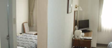 Quarto de hotel com cama, poltrona, escrivaninha e televisão, além de uma janela com cortinas.