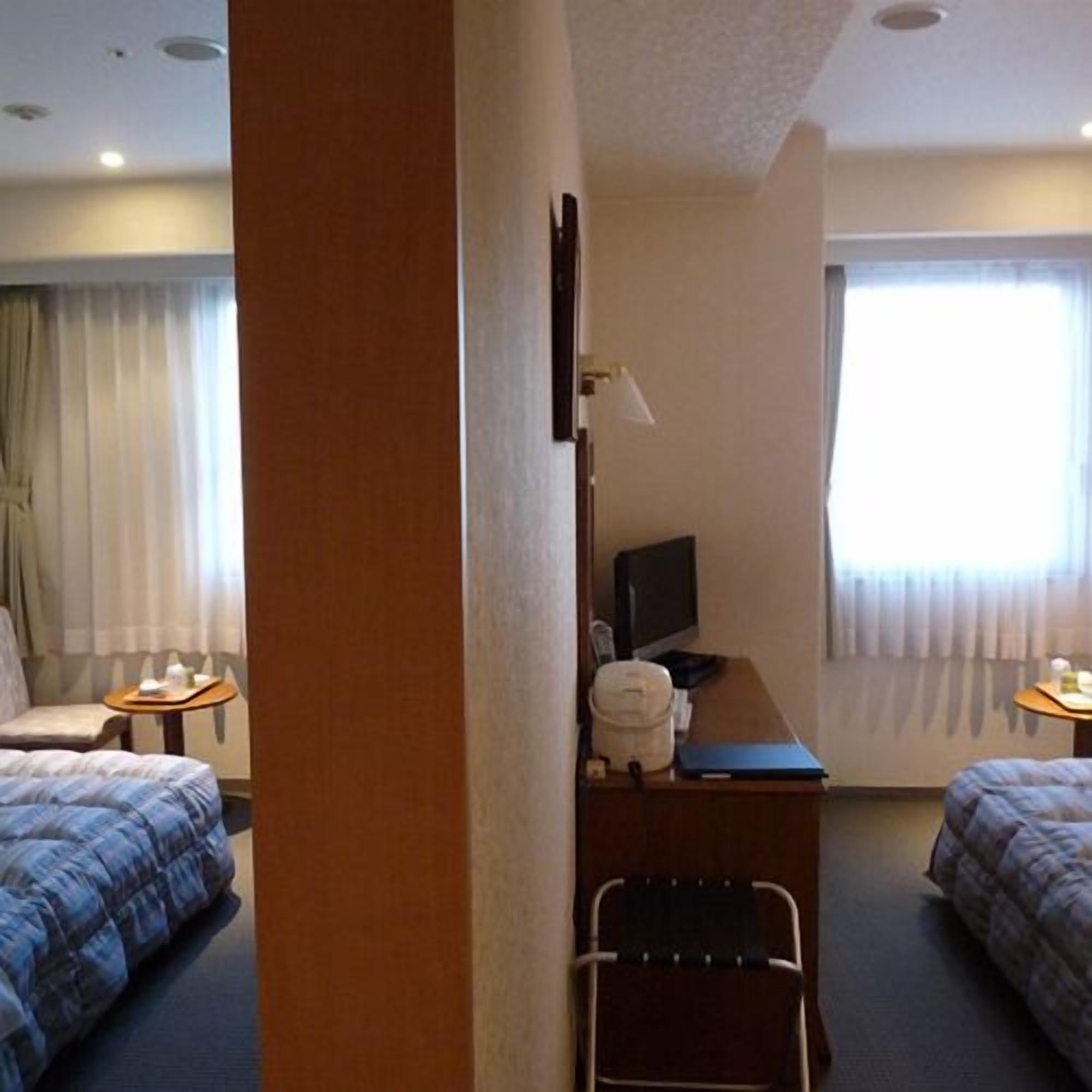Ein Hotelzimmer mit zwei Betten, einem Schreibtisch mit Stuhl, einem Fernseher und einem Fenster mit Vorhängen.