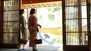 Two individuals in traditional attire walking through a wooden gate into a building with a traditional Japanese architectural style.