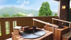 Um deck de madeira com uma banheira de hidromassagem, bancos de madeira e vista para montanhas verdes e um edifício.