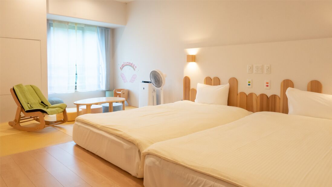 A hotel room with two beds, a rocking chair, a small table, a fan, and a window with curtains.