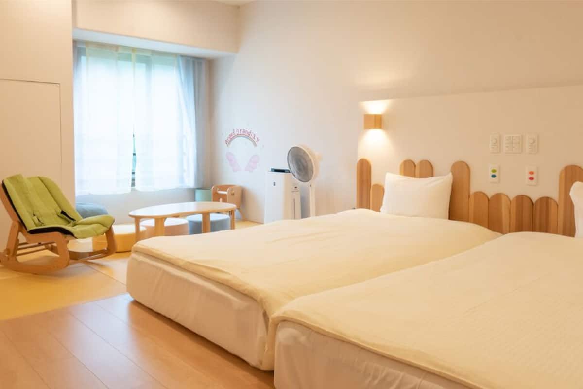 A hotel room with two beds, a rocking chair, a small table, a fan, and a window with curtains.
