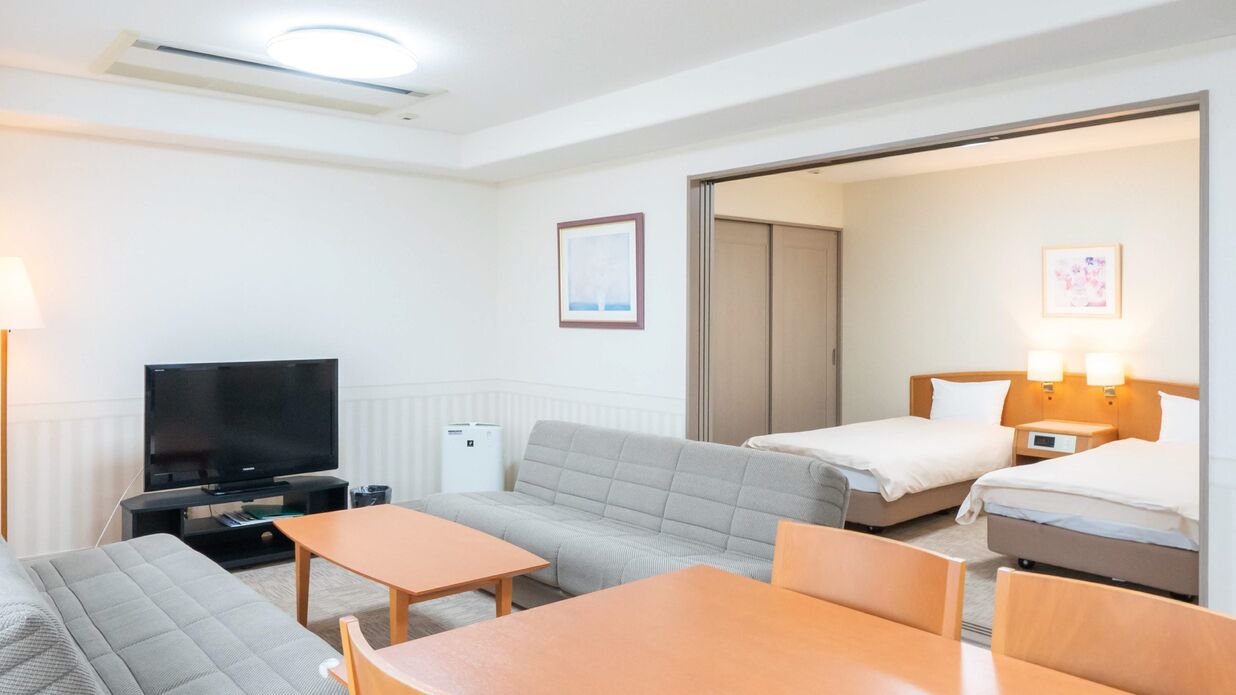 A hotel room with a sofa, two single beds, a TV, a desk, and a lamp.